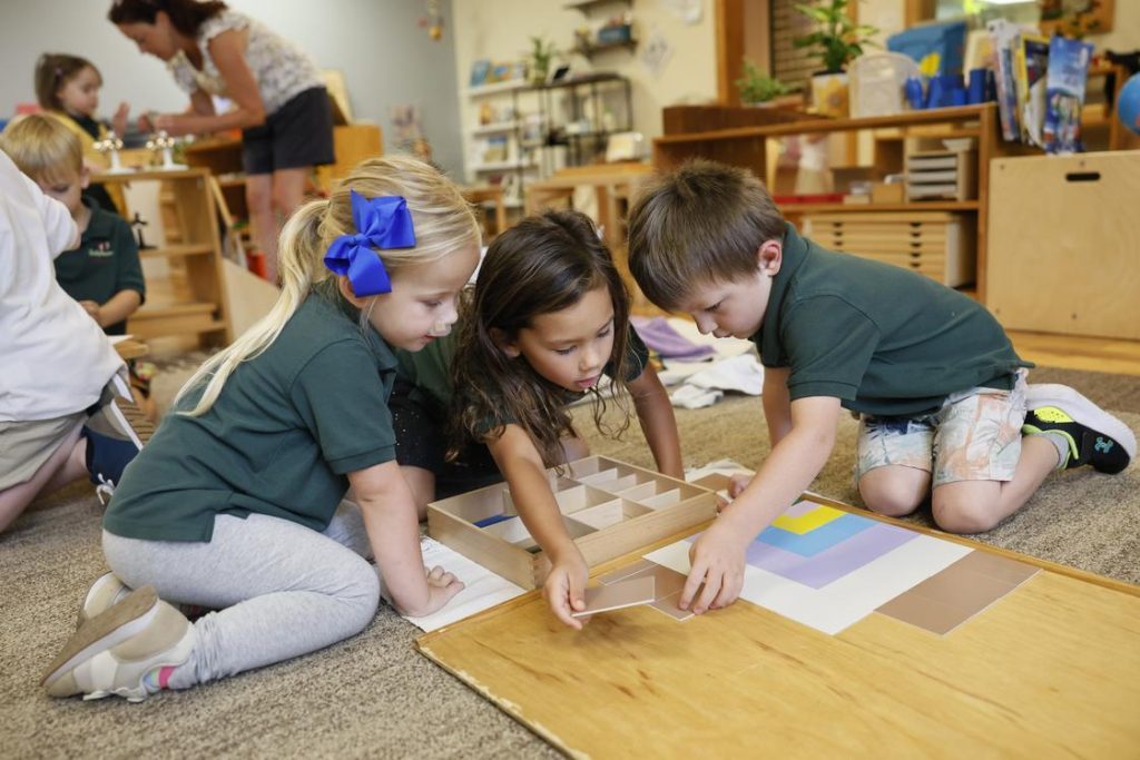 Three children kneel on a classroom carpet, assembling a wooden tile puzzle on the floor.