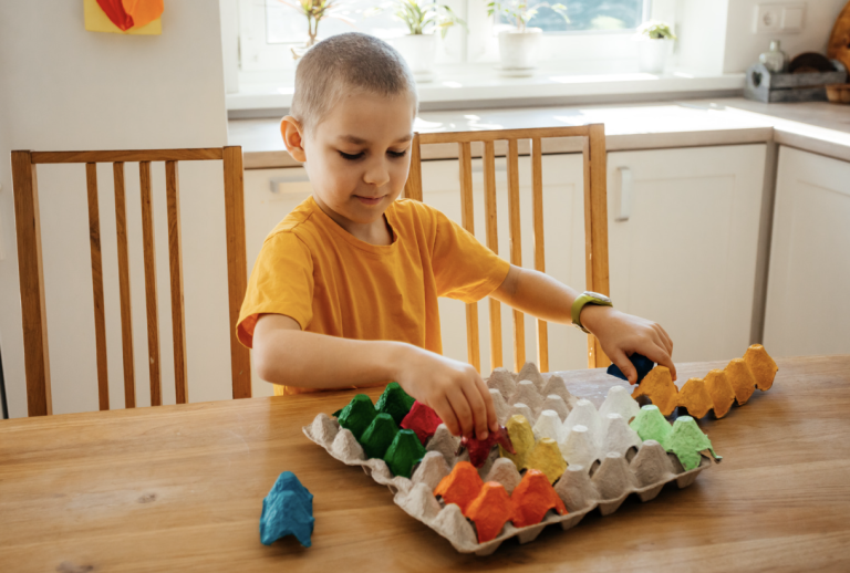 A young boy in an orange shirt stacks colorful paper egg shapes into a gray egg carton on a kitchen table.