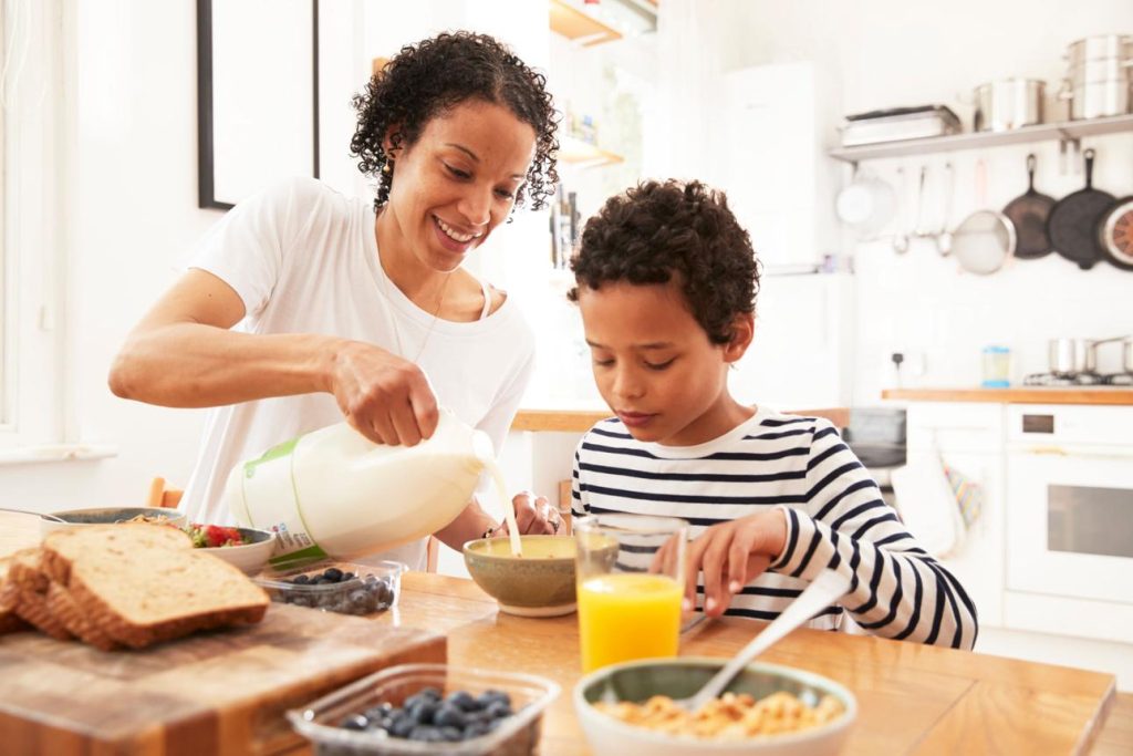Mother and child sharing breakfast, pouring milk into a bowl at a bright kitchen table with toast and blueberries nearby.