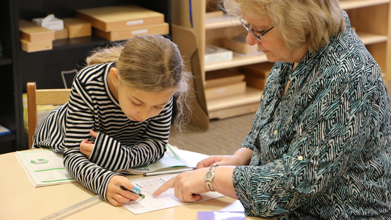 Teacher and student work together at a table, the student writes while the teacher points at a worksheet in a classroom setting
